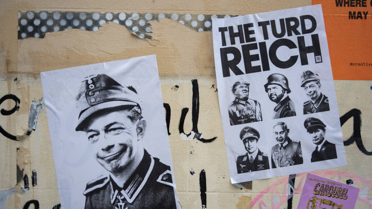 Wand mit verschiedenen aufgeklebten Postern und Graffiti. Im Vordergrund links ist ein Schwarz-Weiß-Porträt von Elon Musk in Militäruniform mit einem Grinsen zu sehen. Daneben rechts befindet sich ein weiteres Poster mit der Überschrift "THE TURD REICH", darunter sind mehrere Porträts von Männern in ähnlichen Uniformen abgebildet, unter anderem Trump und JD Vance.
