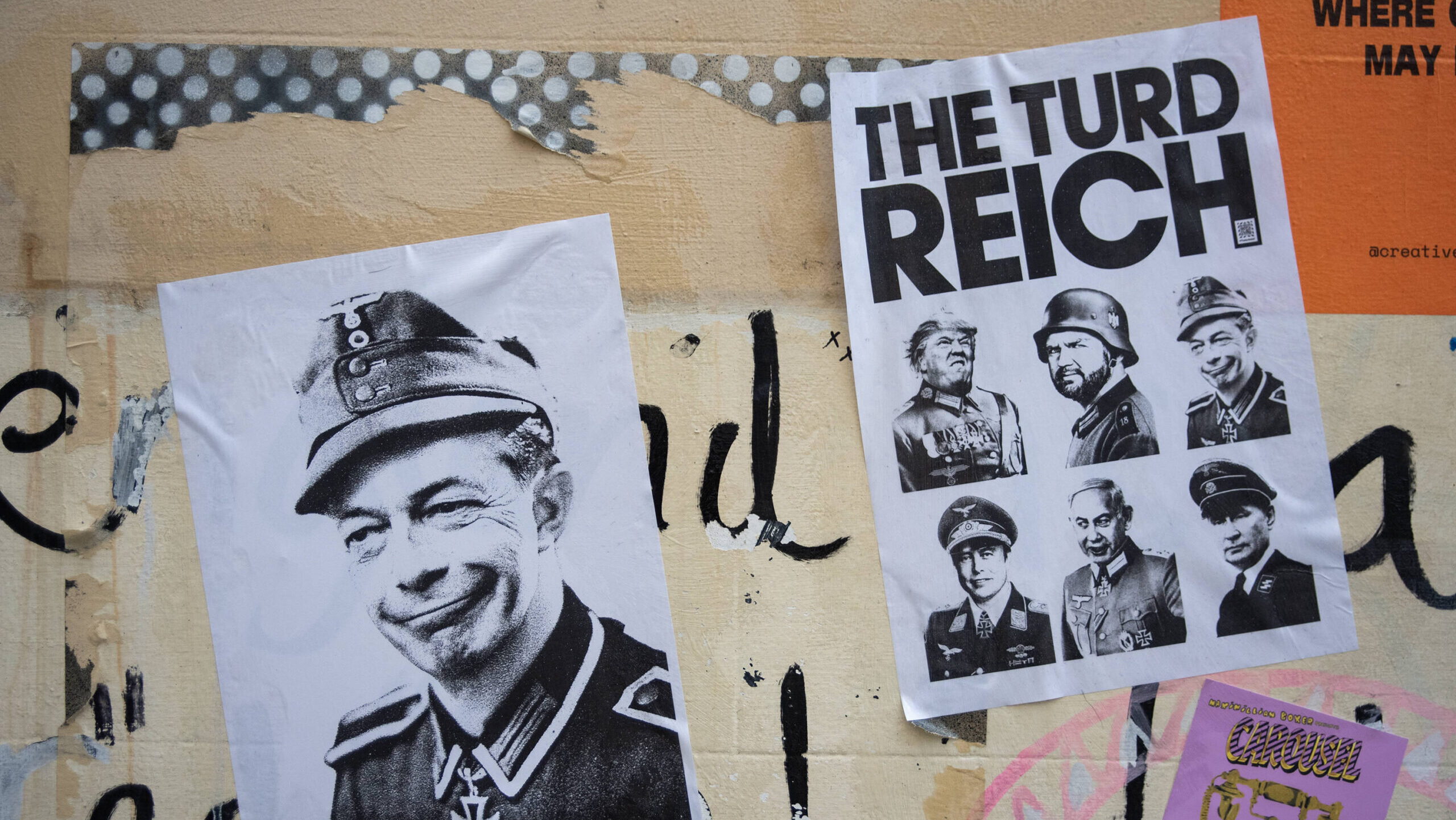 Wand mit verschiedenen aufgeklebten Postern und Graffiti. Im Vordergrund links ist ein Schwarz-Weiß-Porträt von Elon Musk in Militäruniform mit einem Grinsen zu sehen. Daneben rechts befindet sich ein weiteres Poster mit der Überschrift "THE TURD REICH", darunter sind mehrere Porträts von Männern in ähnlichen Uniformen abgebildet, unter anderem Trump und JD Vance.