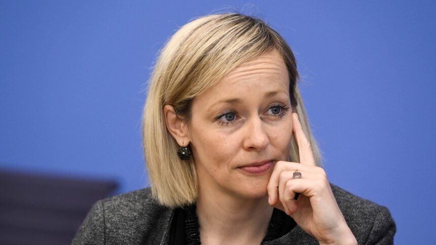 Person mit blondem schulterlangem Haar, die mit der rechten Hand an die rechte Gesichtshälfte fasst, trägt dunkle Kleidung vor blauem Hintergrund.
