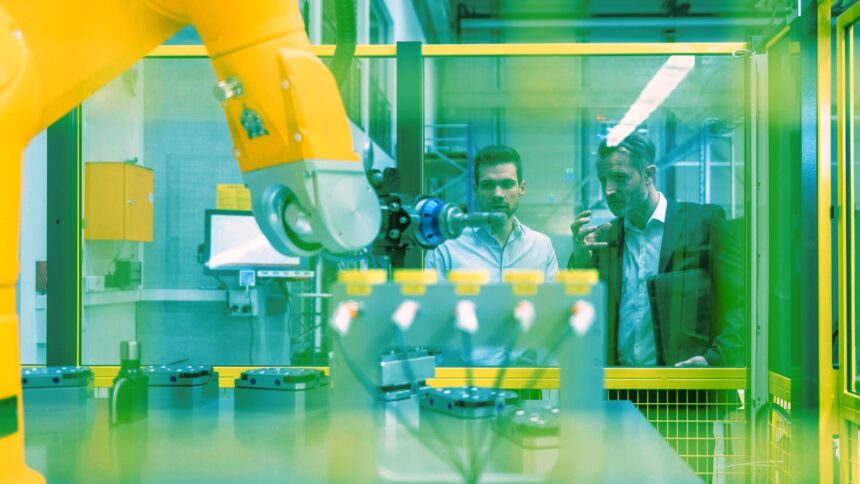 Zwei Personen stehen hinter einer Sicherheitsglasscheibe und blicken auf einen Industrieroboter.