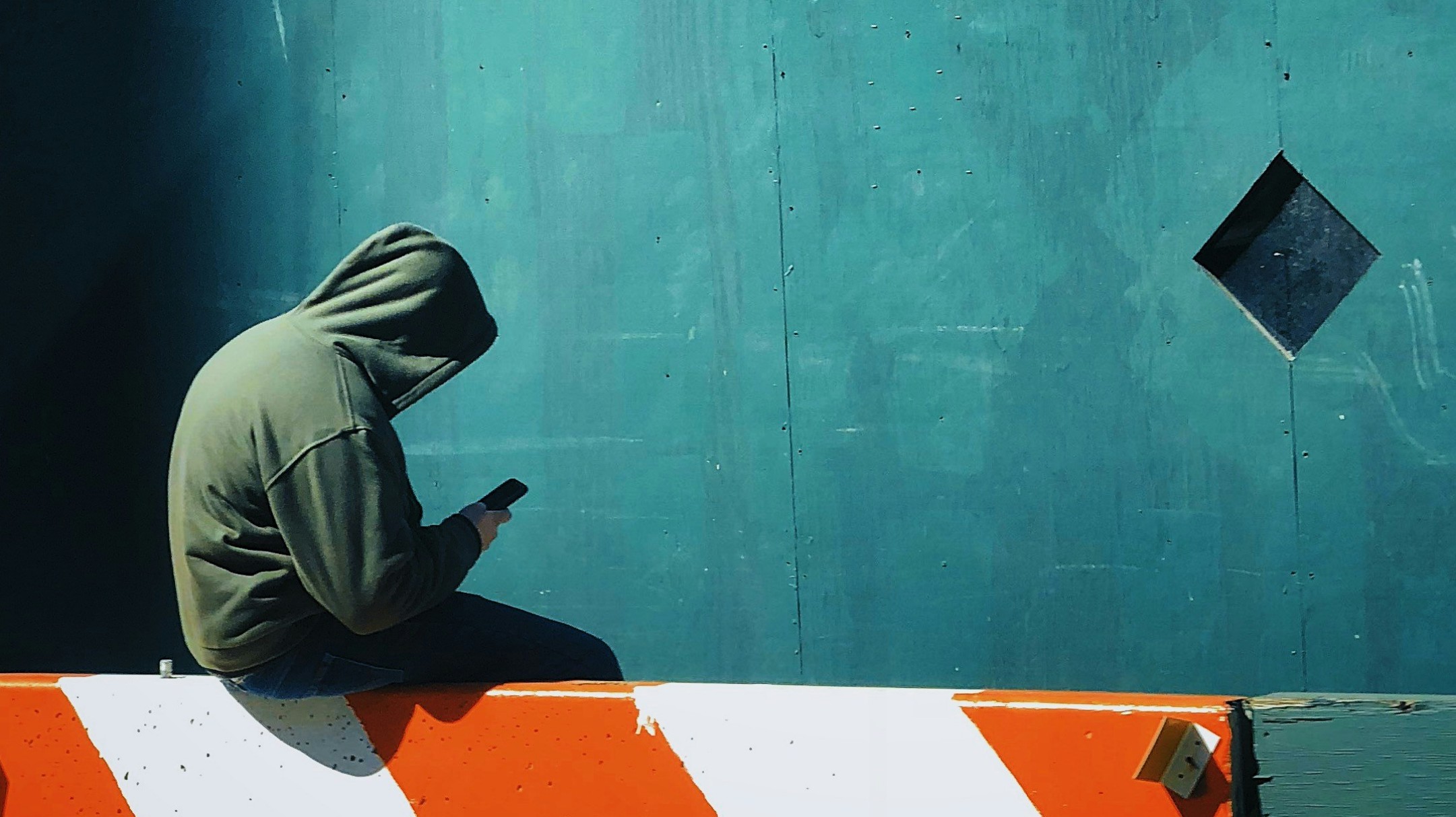 Eine Person im Kaputzenpulli sitzt mit dem Rücken zum Betrachter und schaut aufs Smartphone.
