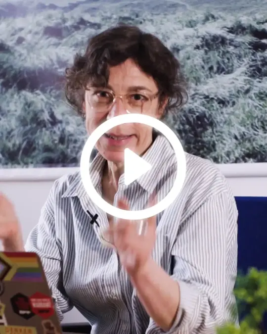 Person sitzt an einem Schreibtisch und schaut mit aktivierenden offenen Händn in die Kamera. Über dem Bild liegt ein Play-Button um zu zeigen, dass es sich um ein Video handelt was durch klicken abgespielt werden kann.