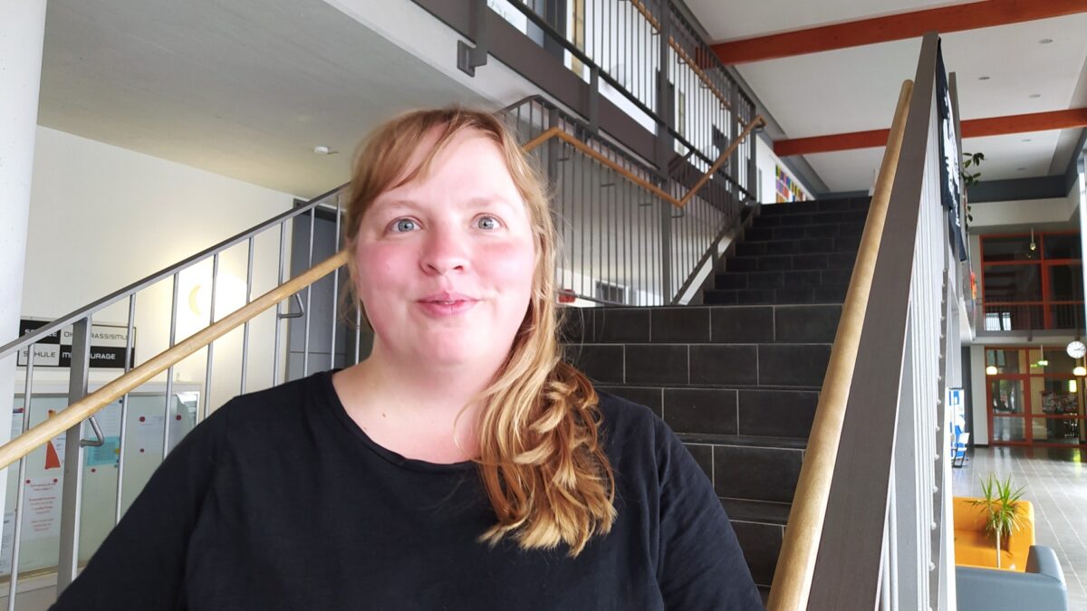 Eine Person mit langen Haaren steht vor einer Treppe in einem Schulgebäude