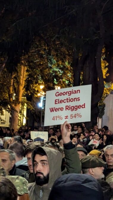 Viele Menschen in einer Stadt bei Nacht, eine Person hält ein Schild hoch auf dem auf Englich steht, dass die Wahl gefälscht gewesen sei.