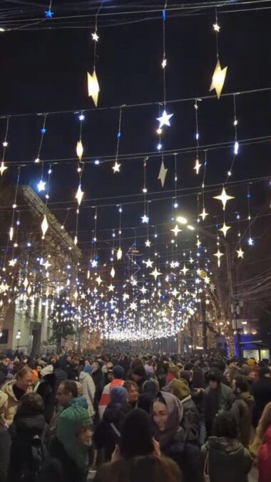 Viele Menschen in einer Straße bei Nacht, über ihnen hängt Weihnachtsschmuck der zwischen den Häusern aufgespannt wurde