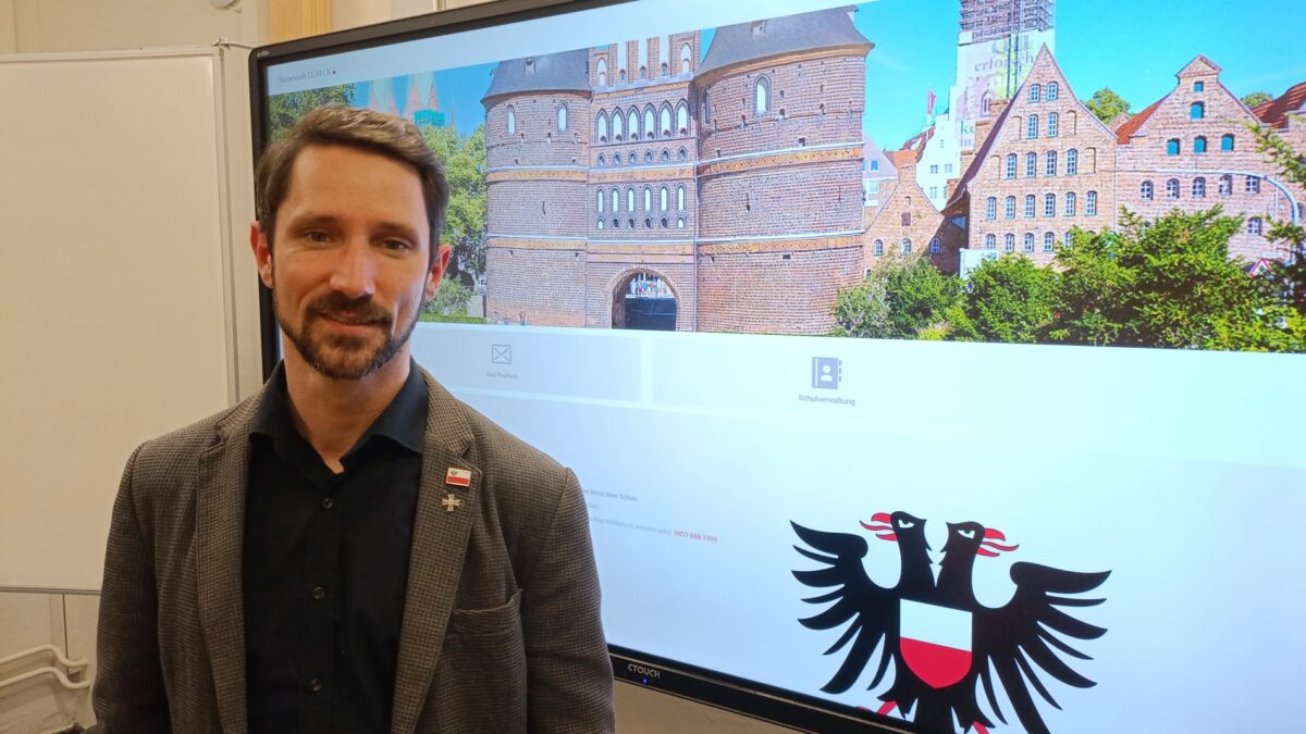Ein Mensch mit Bart und Sakko vor einem Bildschirm, der ein Stadttor zeigt.
