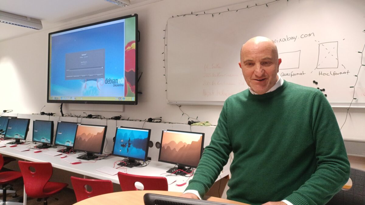 Ein Mensch im grünen Pulli steht vor einem Monitor in einem Computerraum, im Hintergrund sind weitere Monitore