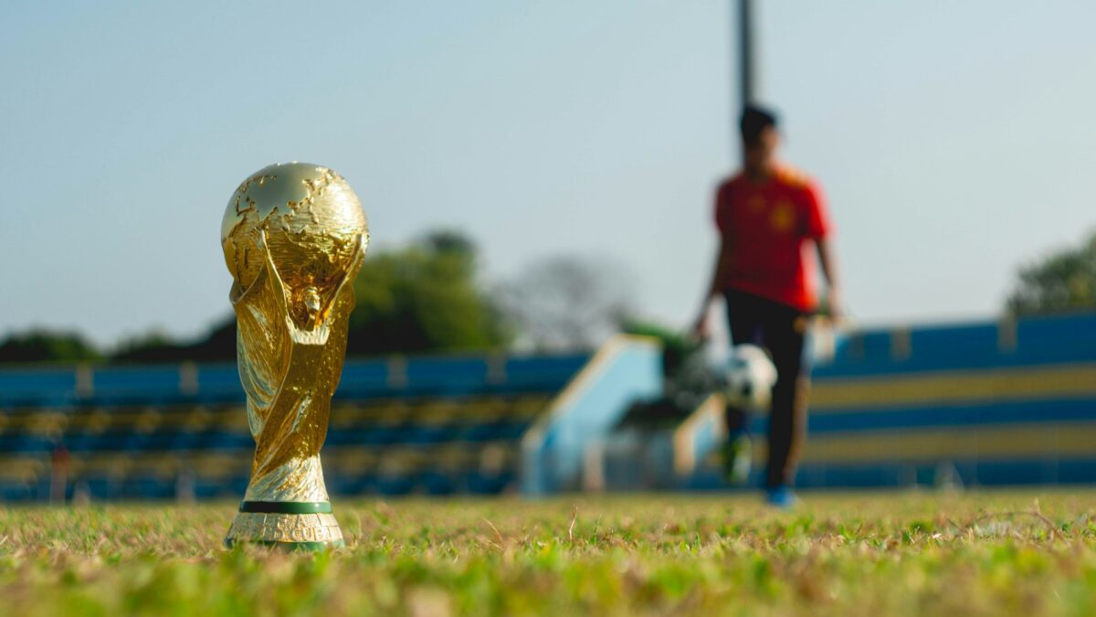 Ein goldener Pokal auf grünem Rasen, im Hintergrund ein Spieler mit Ball