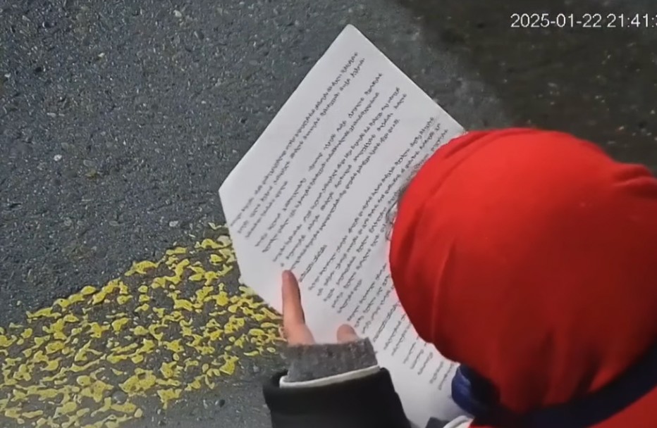 Eine Person mit roter Kapuze und von hinten gefilmt liest einen Text von einem Blatt Papier