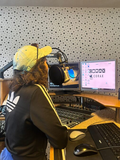 Eine Peson im Sweatshirt und Kappe steht an einem Radio-Arbeitsplatz und spricht ins Mikrofon.