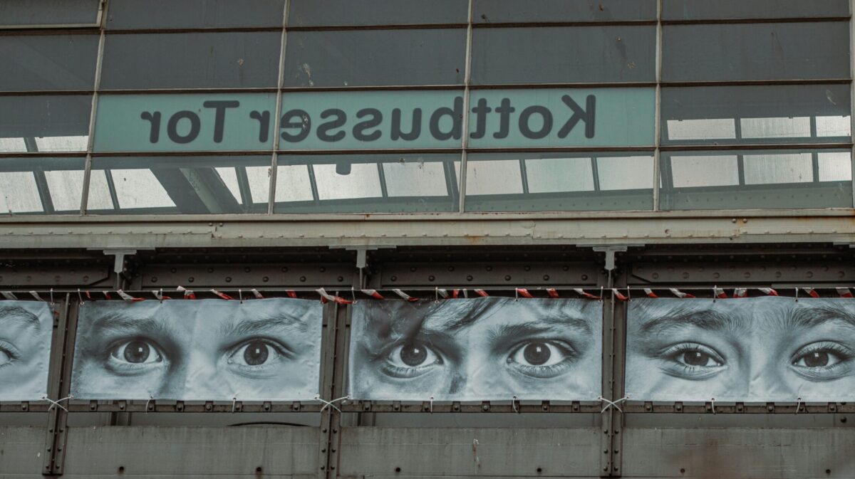 Eine Installlation mit Postern auf denen Augenpaare abgedruckt sind und die an der Hochbahn-Station Kottbusser Tor hängen, Schriftzug scheint spiegelverkehrt durchs Glas.