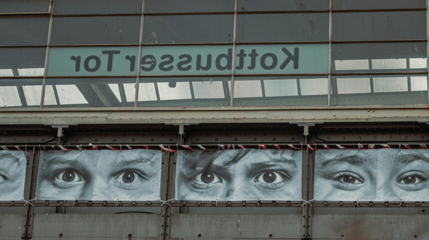 Eine Installlation mit Postern auf denen Augenpaare abgedruckt sind und die an der Hochbahn-Station Kottbusser Tor hängen, Schriftzug scheint spiegelverkehrt durchs Glas.