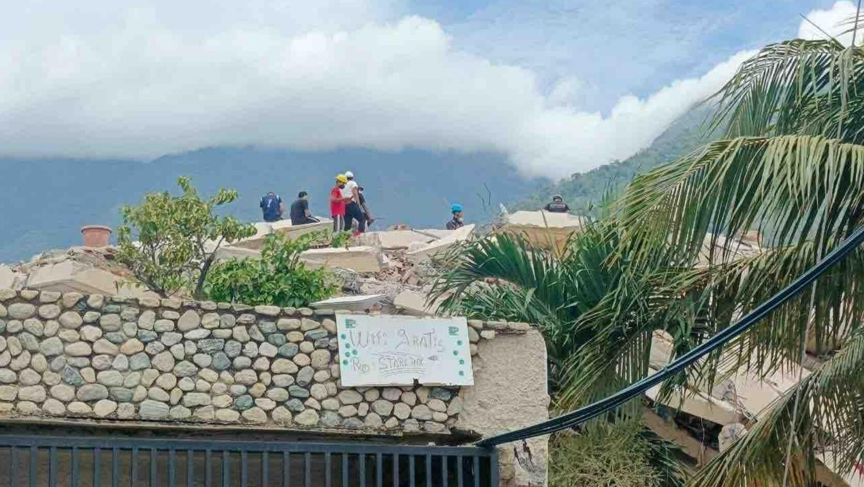 Ein leicht unscharfes Foto in einem bergigen Dorf, im Vordergrund ein Schild mit der Aufschrift „Wifi: Gratis“, im Hintergrund Katastrophenhelfer:innen