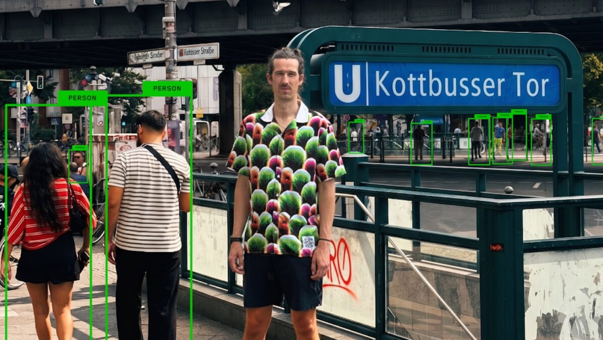 Mann mit kurzärmligem Hemd mit grün-rotem Muster und dunklen Shorts steht vor dem U-Bahnhof Kottbusser Tor in Berlin