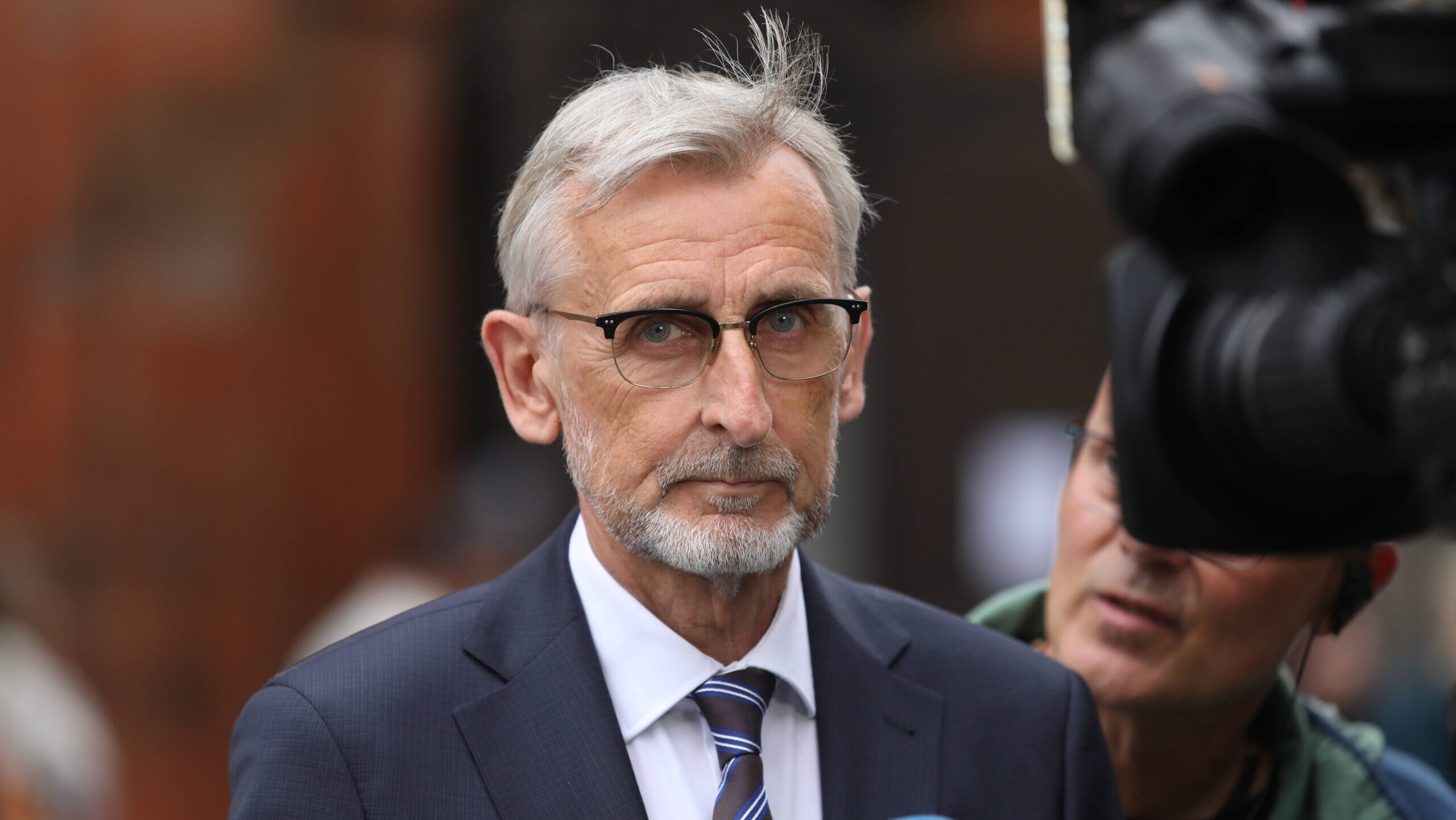 Mann mit grauen Haaren, Anzug und Brille, rechts im Bild eine Kamera