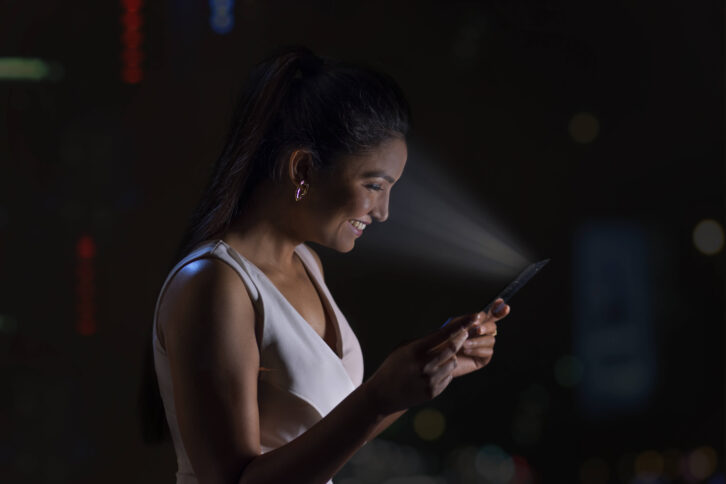 Eine Person schaut lächelnd auf ihr Handy, während das Handylicht auf ihr Gesicht fällt und es in Abgrenzung zur dunklen Umgebung stark beleuchtet.