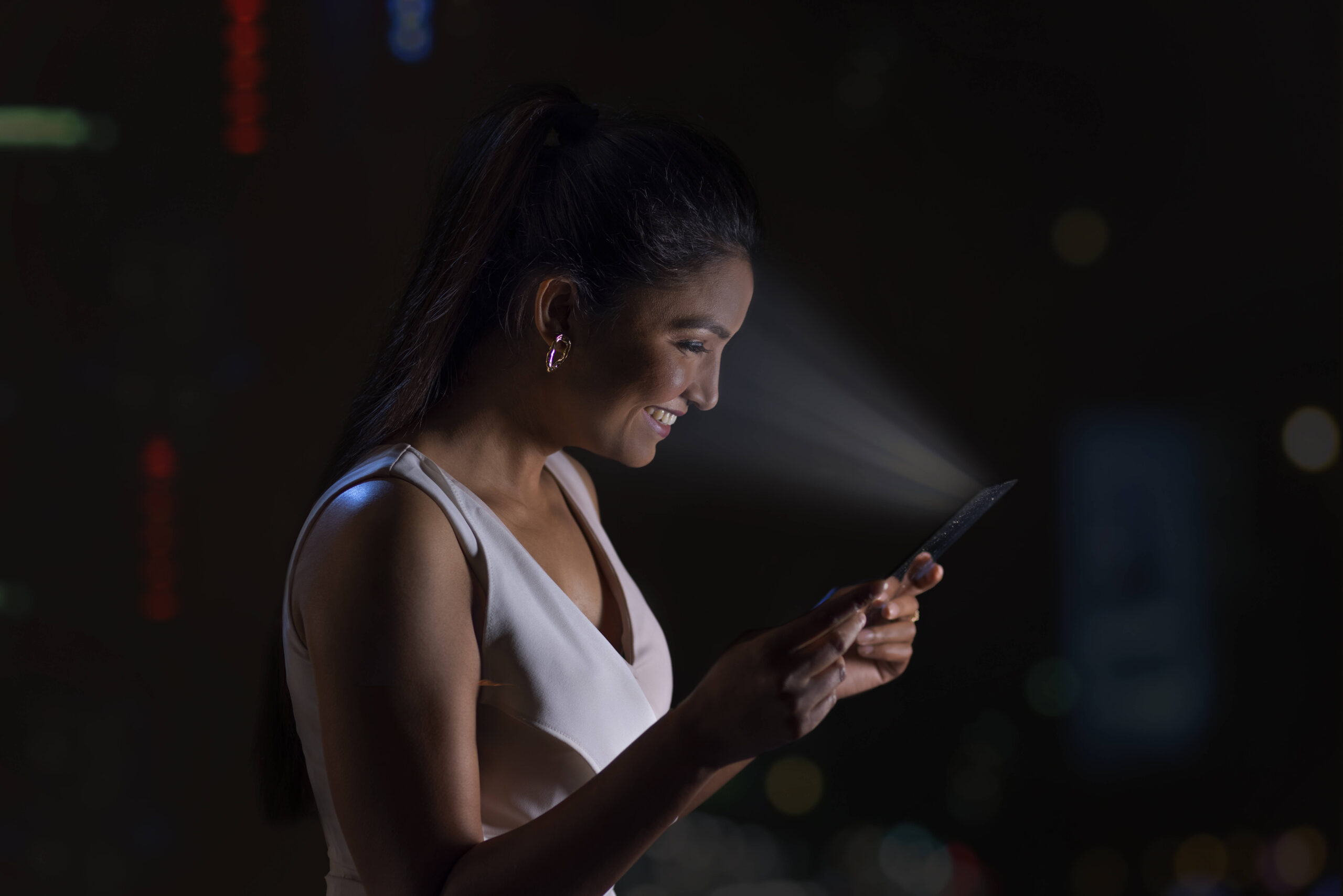 Eine Person schaut lächelnd auf ihr Handy, während das Handylicht auf ihr Gesicht fällt und es in Abgrenzung zur dunklen Umgebung stark beleuchtet.