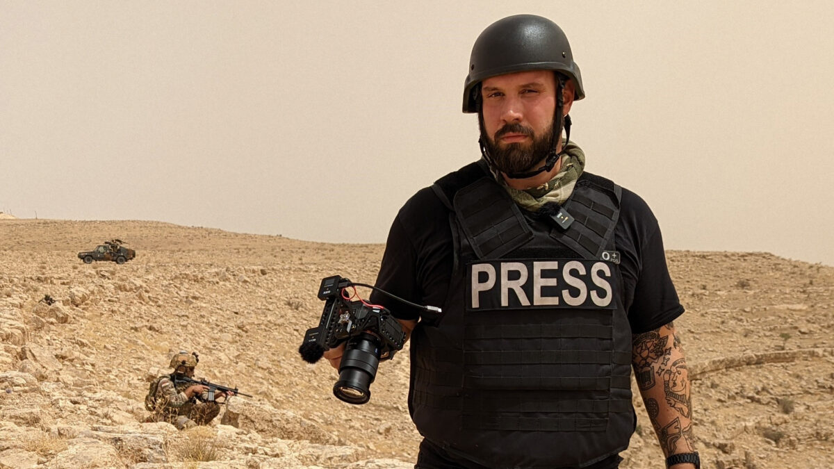 Ein Mensch in Presseweste und mit Kamera steht in einer kargen Landschaft, hinter ihm kniet eine Person mit Sturmgewehr