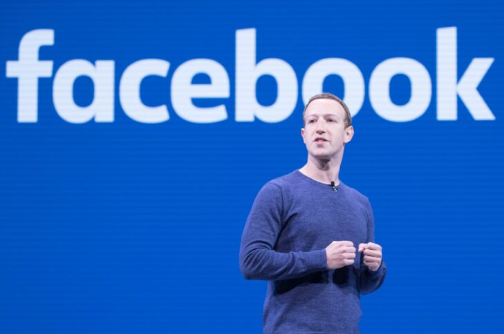 Ein blauer Hintergrund mit einem großen weißen Schriftzug "Facebook", davor ein junger Mark Zuckerberg mit kurzen Haaren im blauen Longsleeve