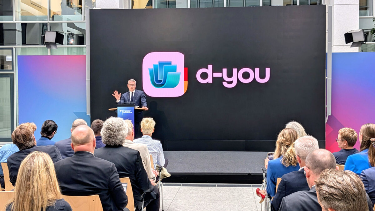 Eine Bühne mit Sprecher:innenpult, dahinter eine Leinwand mit dem Schriftzug "d-you".