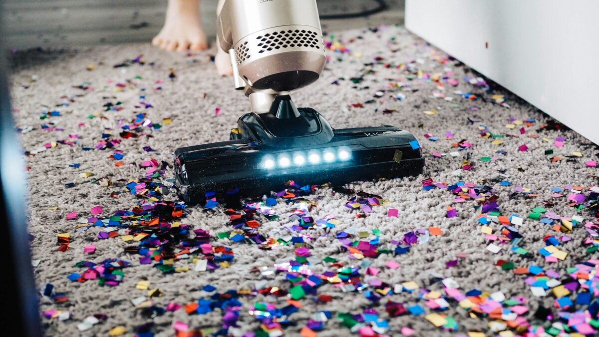 Ein moderner Staubsauger saugt bunte Konfetti-Schnipsel von einem braunen Wollteppich