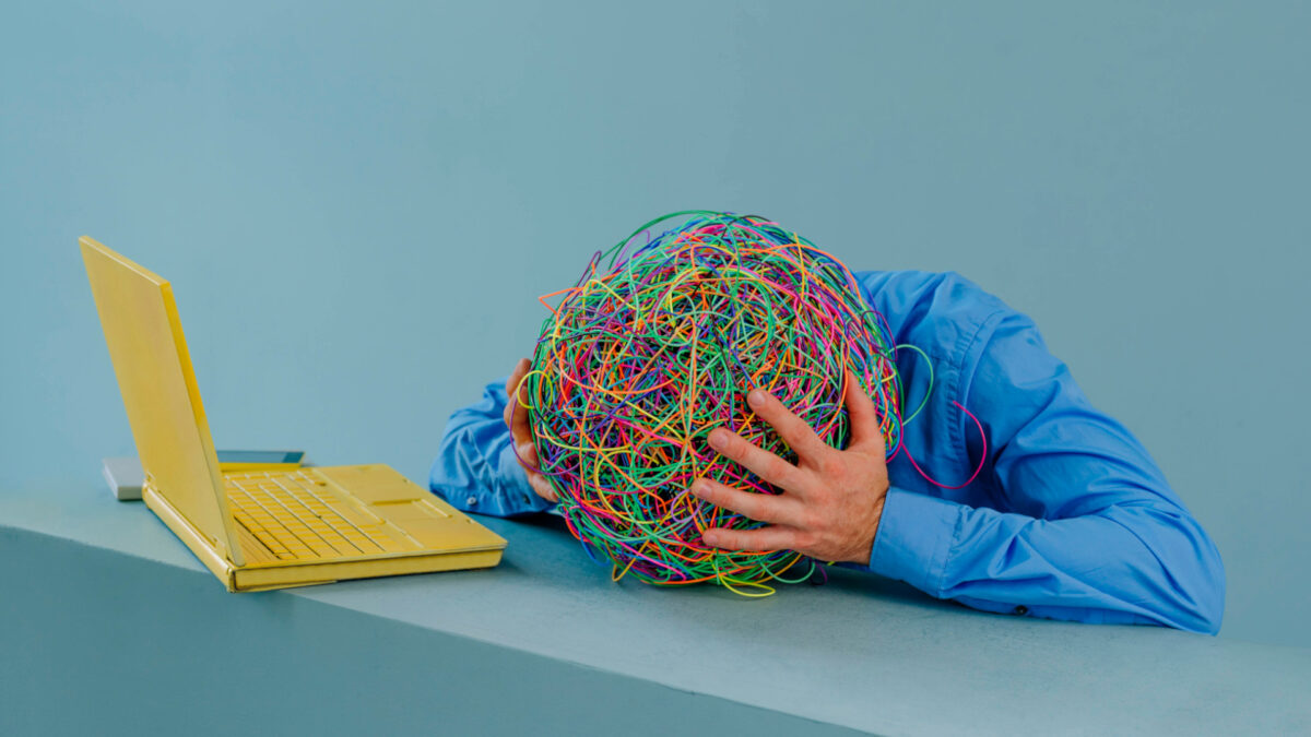 Das Symbolbild zeigt eine Person mit einem Kopf aus bunten Gummiringen. Sie legt den Kopf auf den Schreibtisch und hält ihn fest. Vor ihr ein gelber Laptop.