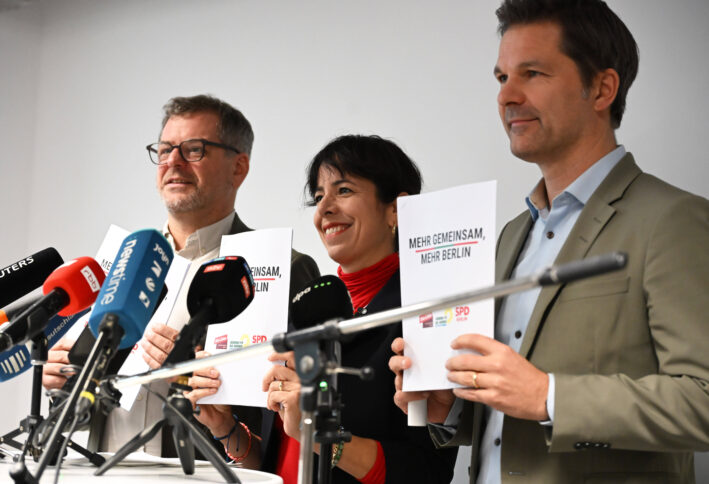 Drei Menschen stehen hinter Mikrofonen und halten Papiere in Kameras