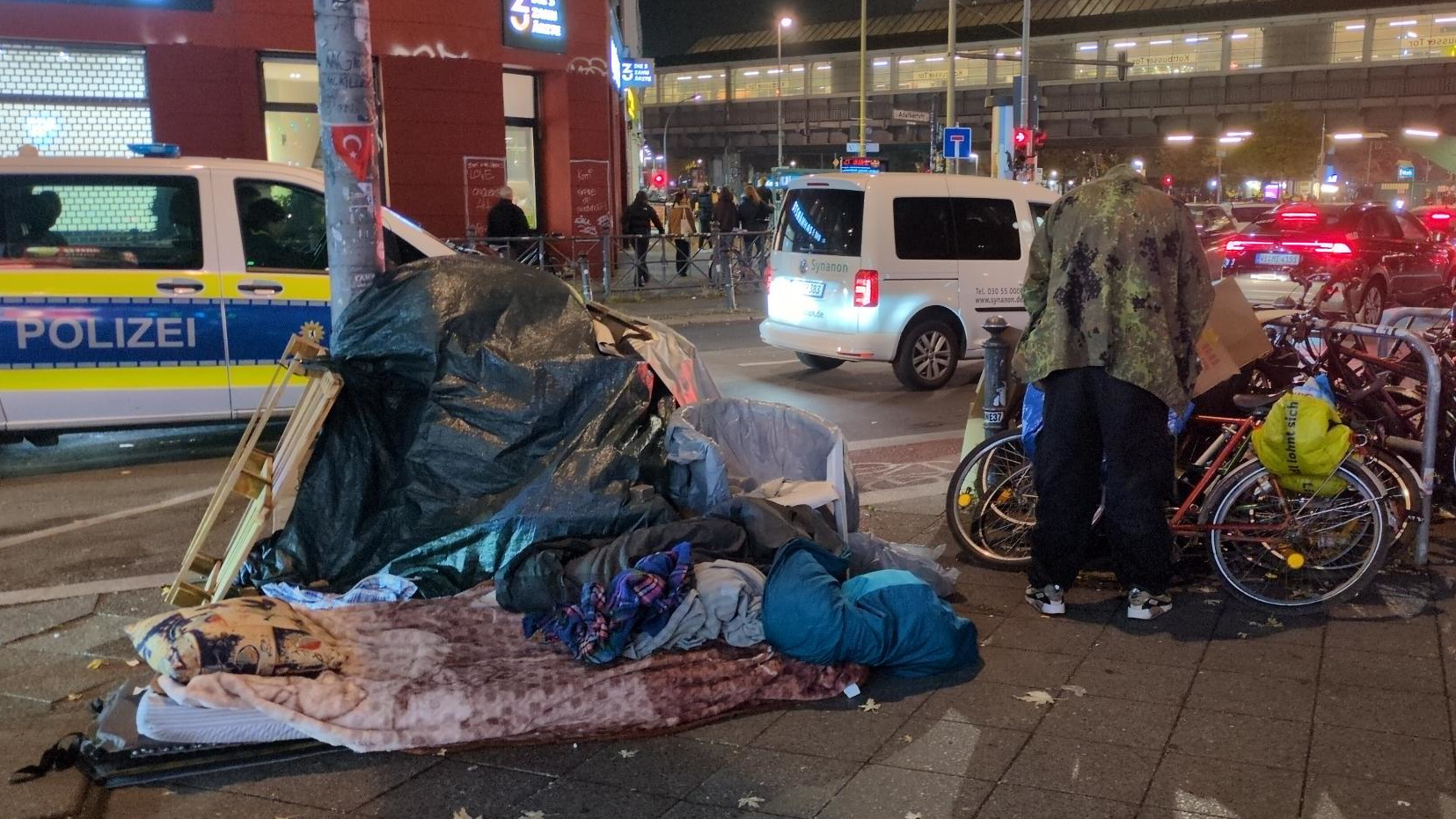 Ein Matratzenlager, ein Mann in Tarnfleck-Jacke steht gebückt dahinter und ist von hinten zu sehen. Am Bildrand kommt ein Polizeitransporter angefahren.