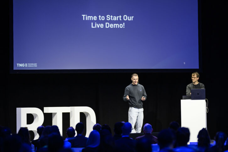 Zwei Menschen stehen auf einer großen Vortragsbühne, auf der Slide hinter ihnen steht "Time to start our live demo"